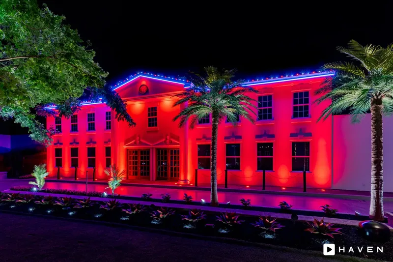 A large two-story building washed in red light with blue roofline lighting, palm trees and lit landscaping.