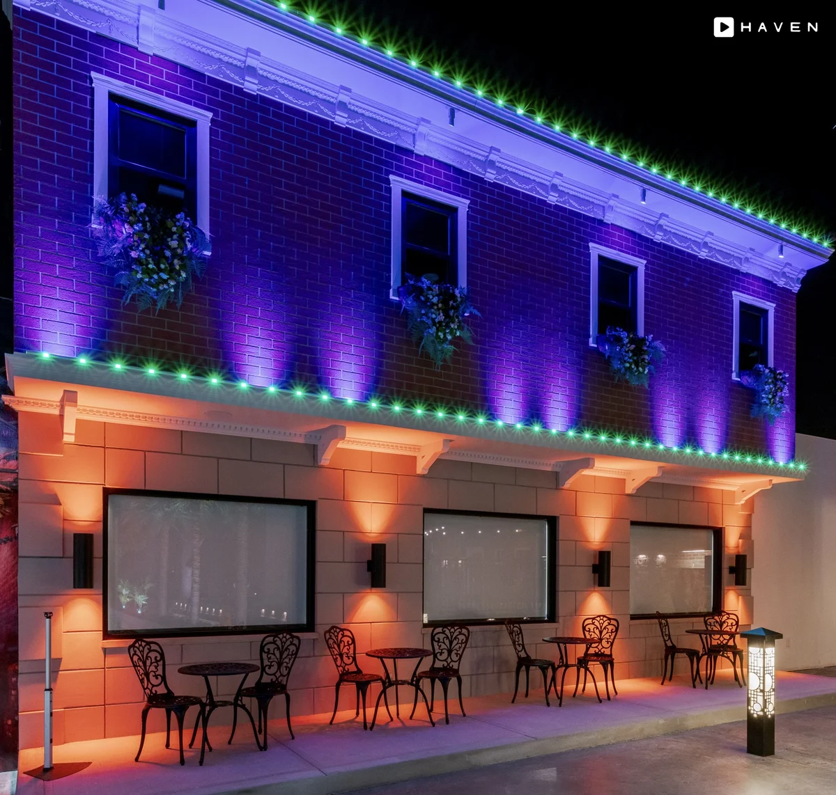 A two-story brick storefront at night, washed in purple light with green permanent lights along both rooflines and warm orange lights over the patio tables.