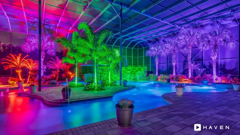A screened pool enclosure with palm trees uplit in red, pink, green and purple around a glowing blue pool.