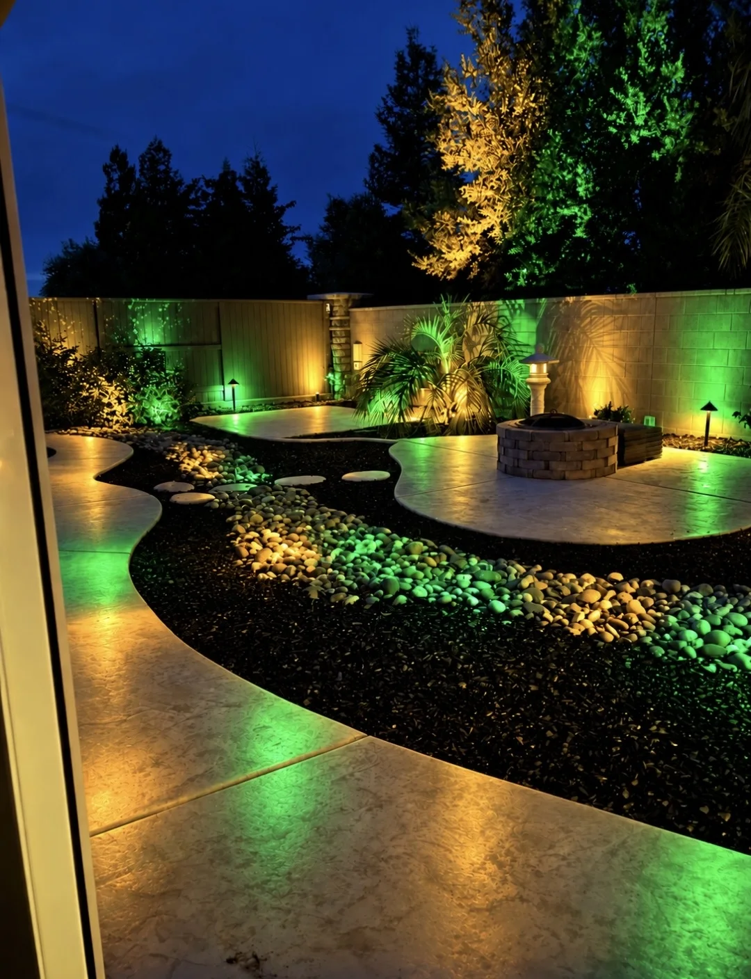 A backyard at night with a fire pit, a curving lit walkway, glowing river rock and a fence and trees uplit in warm white and green.