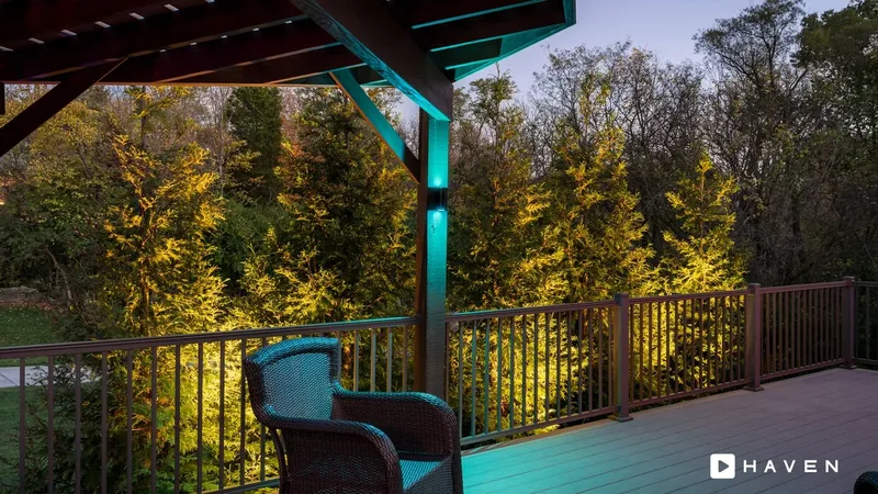 A deck under a pergola lit in teal, looking out at evergreen trees uplit in warm white.