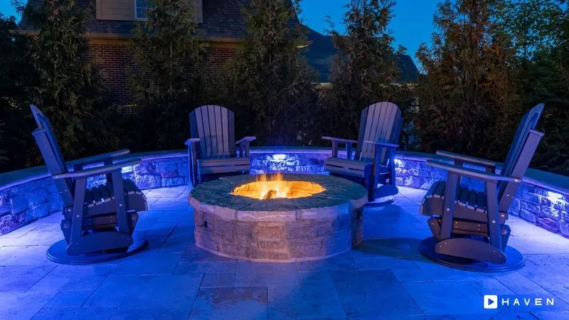 A round stone fire pit with four rocking chairs, inside a curved stone seat wall lit in blue.