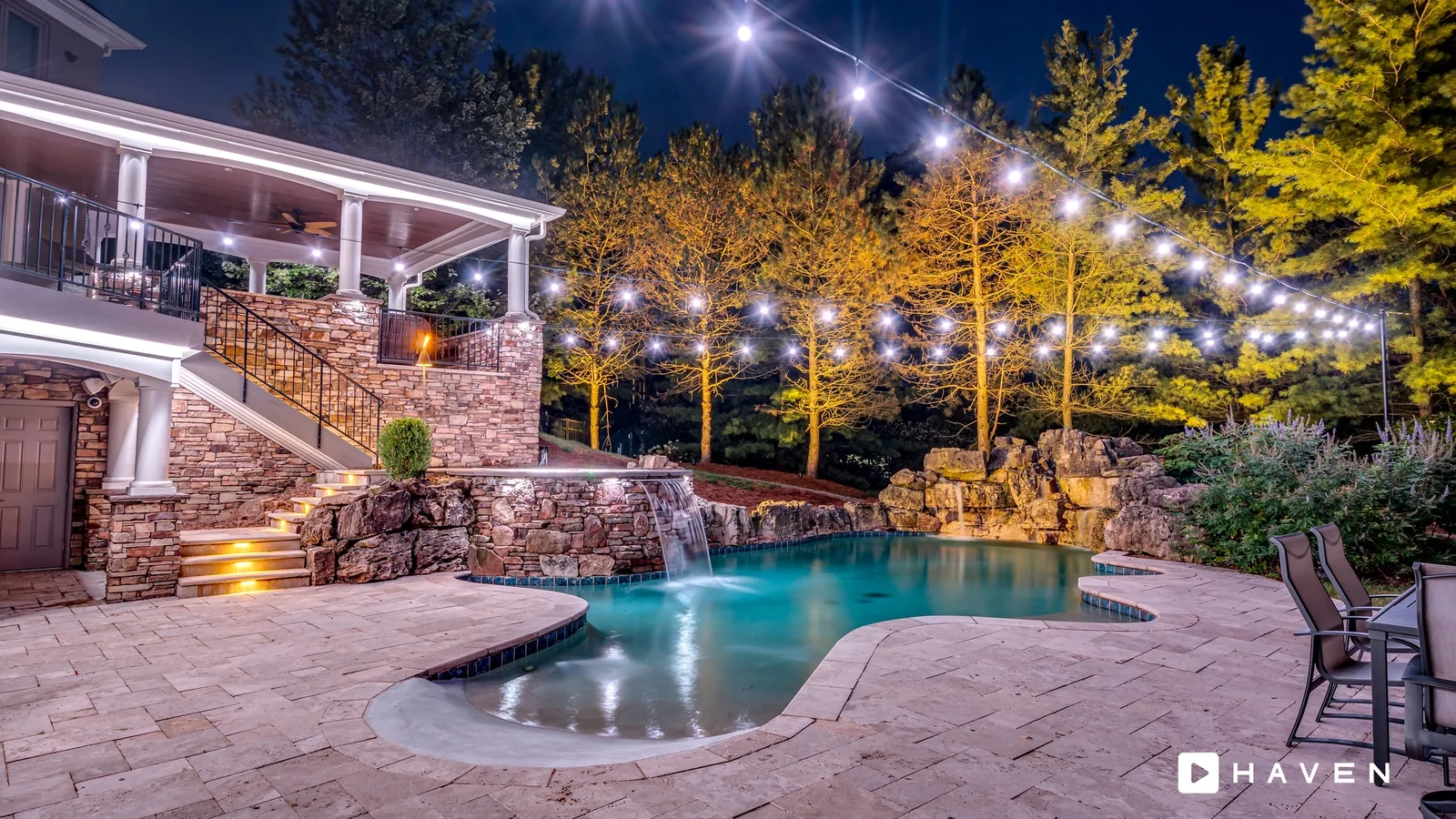 A backyard pool with a stone waterfall, lit steps and a covered porch, under string lights, with tall trees uplit in warm white.