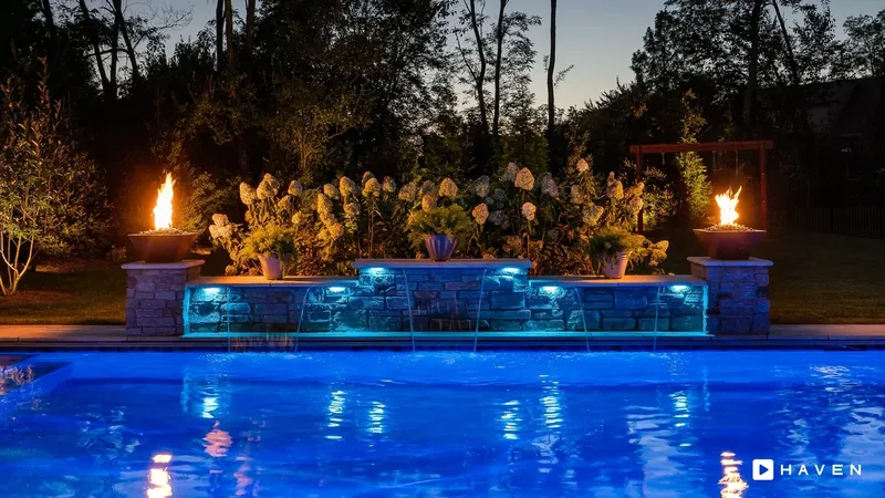 A pool with a lit stone waterfall wall, fire bowls on each side and uplit hydrangeas behind.