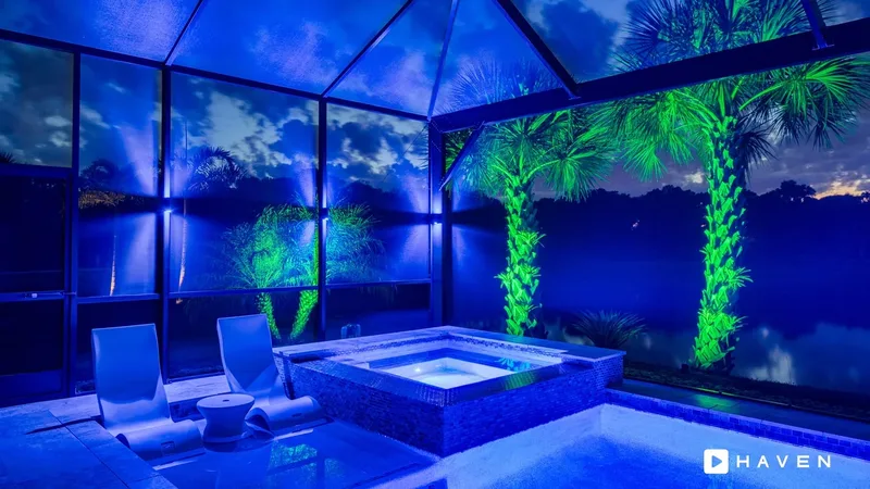 A spa and sun shelf in a screened pool enclosure lit in blue, with palm trees uplit in green.