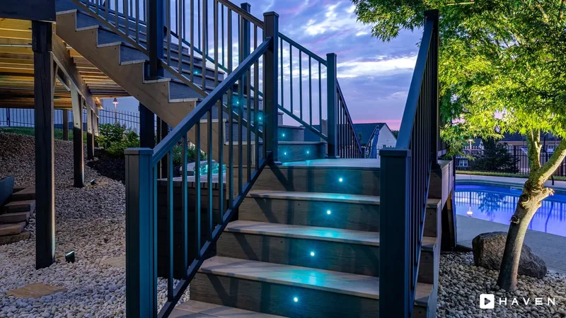 Deck stairs with teal step lights beside a pool and an uplit tree at dusk.