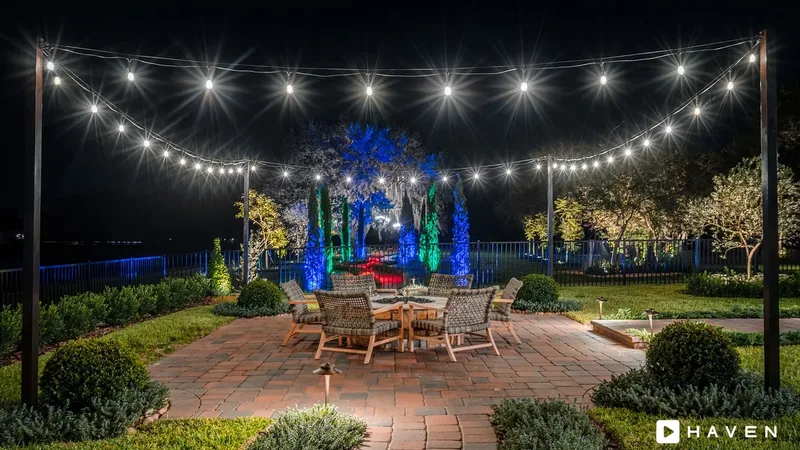 A brick patio with a fire table under string lights, with cypress trees uplit in blue and green beyond the fence.