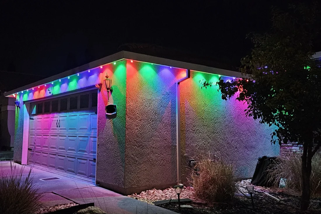 A garage and house wall lit in red, green, blue and purple by permanent lights along the roofline at night.